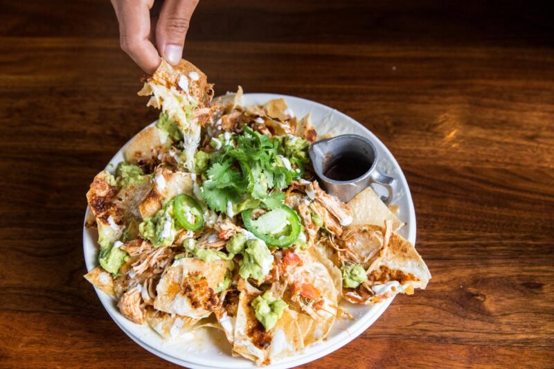 Hand dipping into a cheesy plate of Hub 51's Famous Pulled Chicken Nachos 