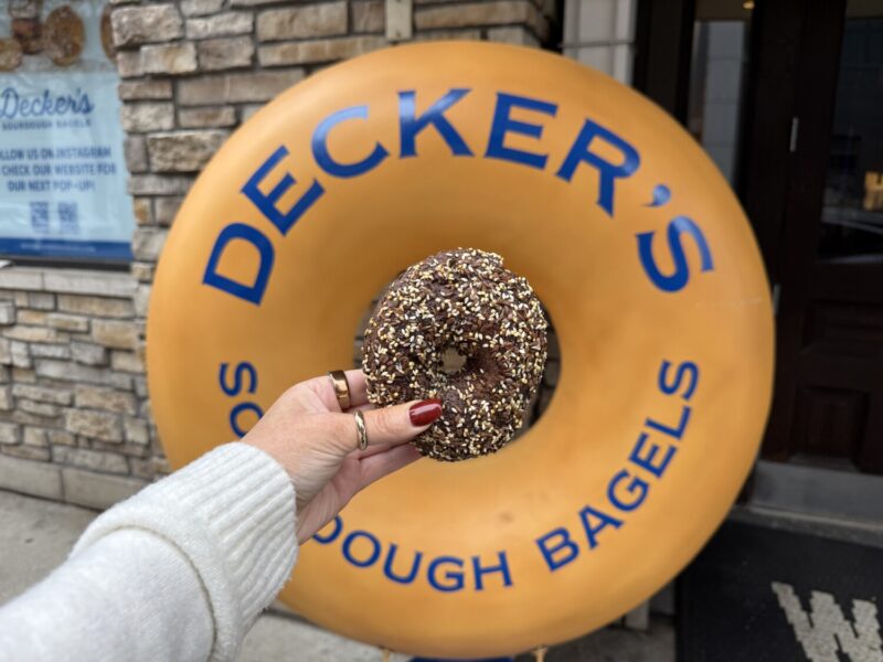 A hand holding up a bagel in the center of a large bagel shaped sign reading Decker's Sourdough Bagels