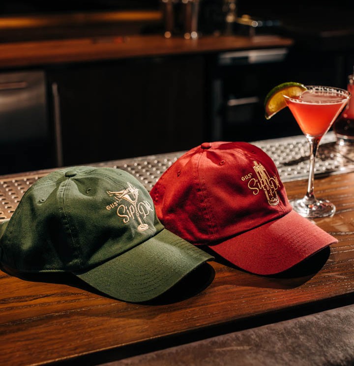 A green dad hat and red dad hat with the Gus' Sip & Dip logos on a bar next to a Cosmopolitan cocktail