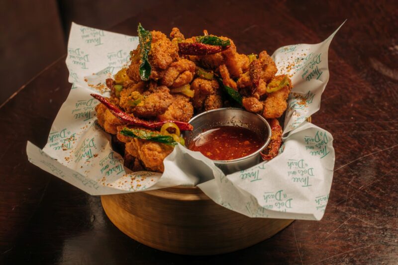 Three Dots and dash firecracker popcorn chicken with Szechuan Pepper & Dried Chilis, Garlic Oil, Red Curry Hot Honey in a white paper lined bowl with sauce