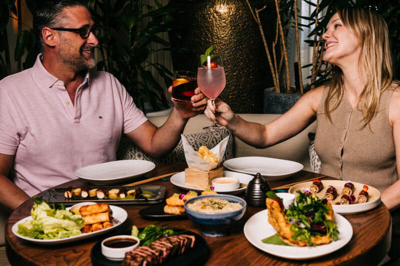 Two people toasting with cocktails and enjoying a spread of food items at Miru