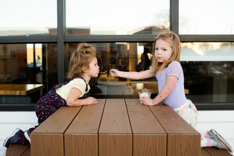 Two kids seating at a bench, with one of them feeding the other with a spoon