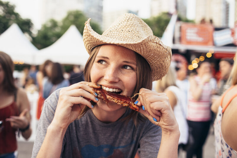 A woman eating a rib at a festival with a cowboy hat on