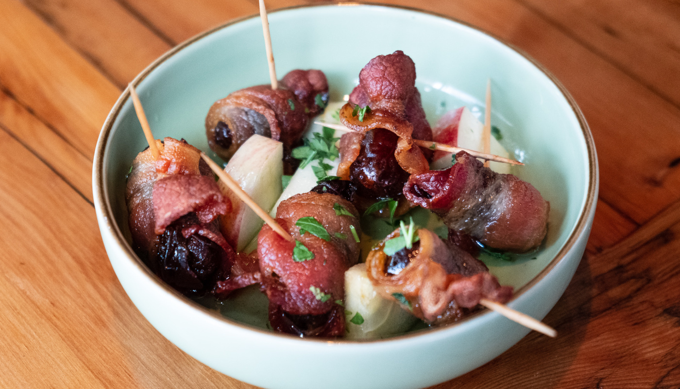 A bowl of bacon-wrapped dates, apple chunks and garnish, with $1 from each order donated to Kitchen Possible
