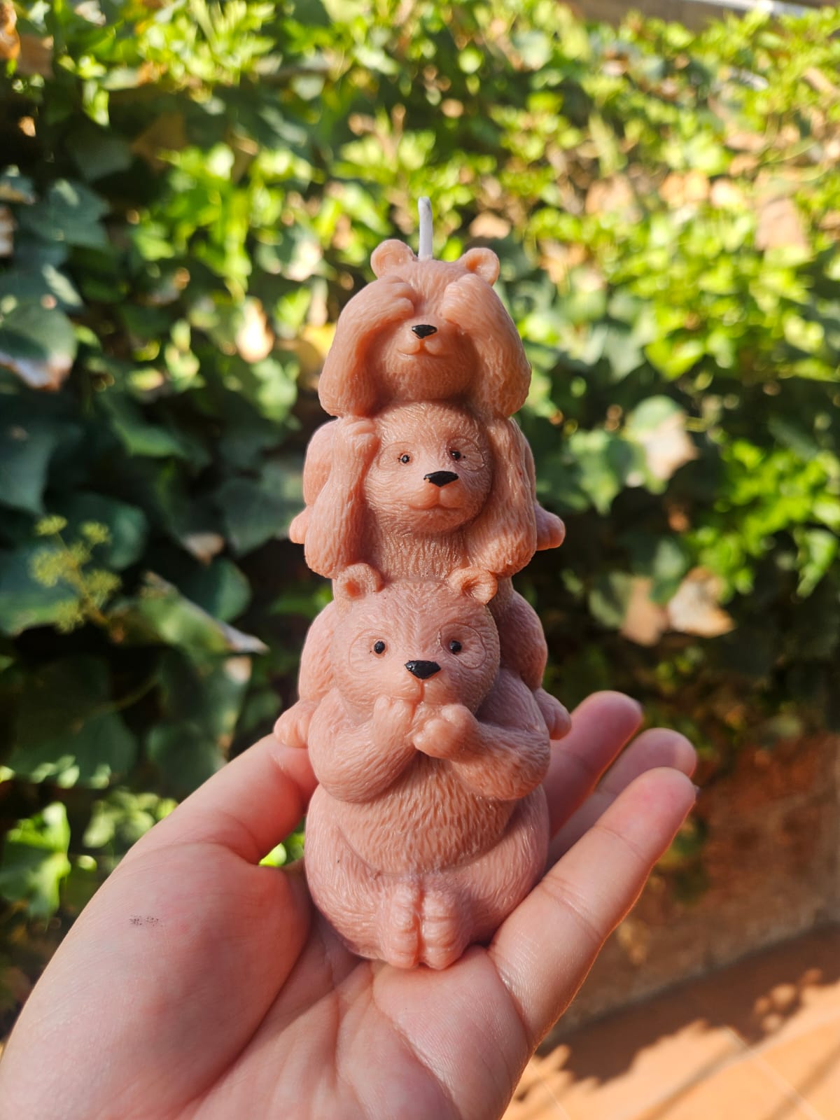 Vela artesanal de soya con tres ositos, sostenida en una mano frente a un jardín