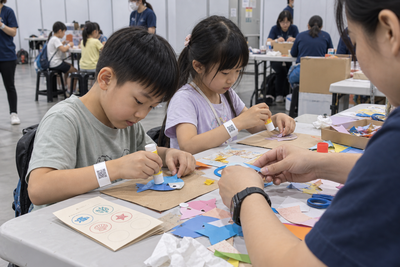 AI 情境示意：孩子一起製作紙魚，工作人員協助，桌邊留有紙卡、手腕配戴 QR 手環，非現場實拍