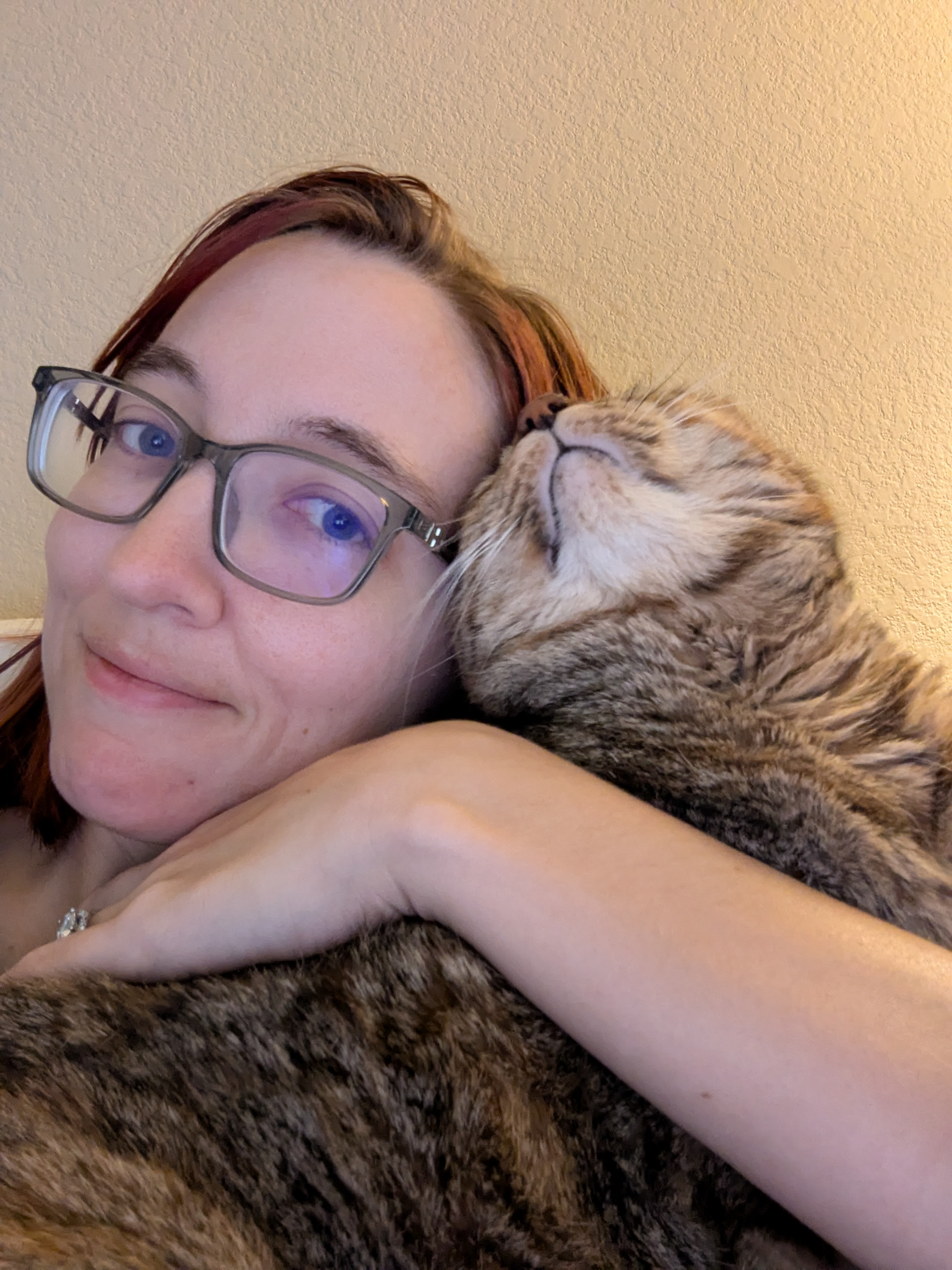Person with glasses smiling as a tabby cat enthusiastically smooshes its face into their cheek.