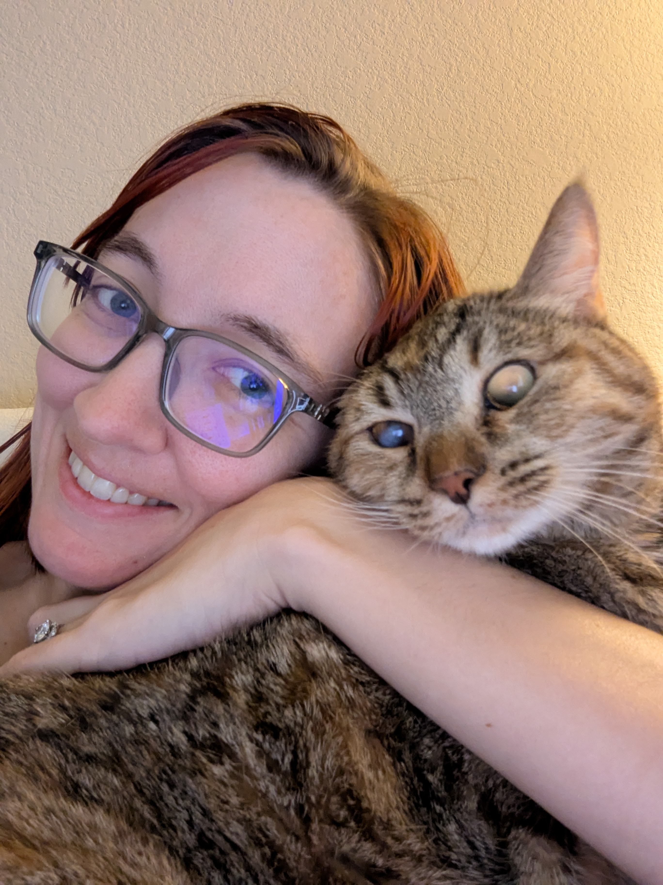Person with glasses smiling while holding a tabby cat resting its squished face on their arm, looking wide-eyed.