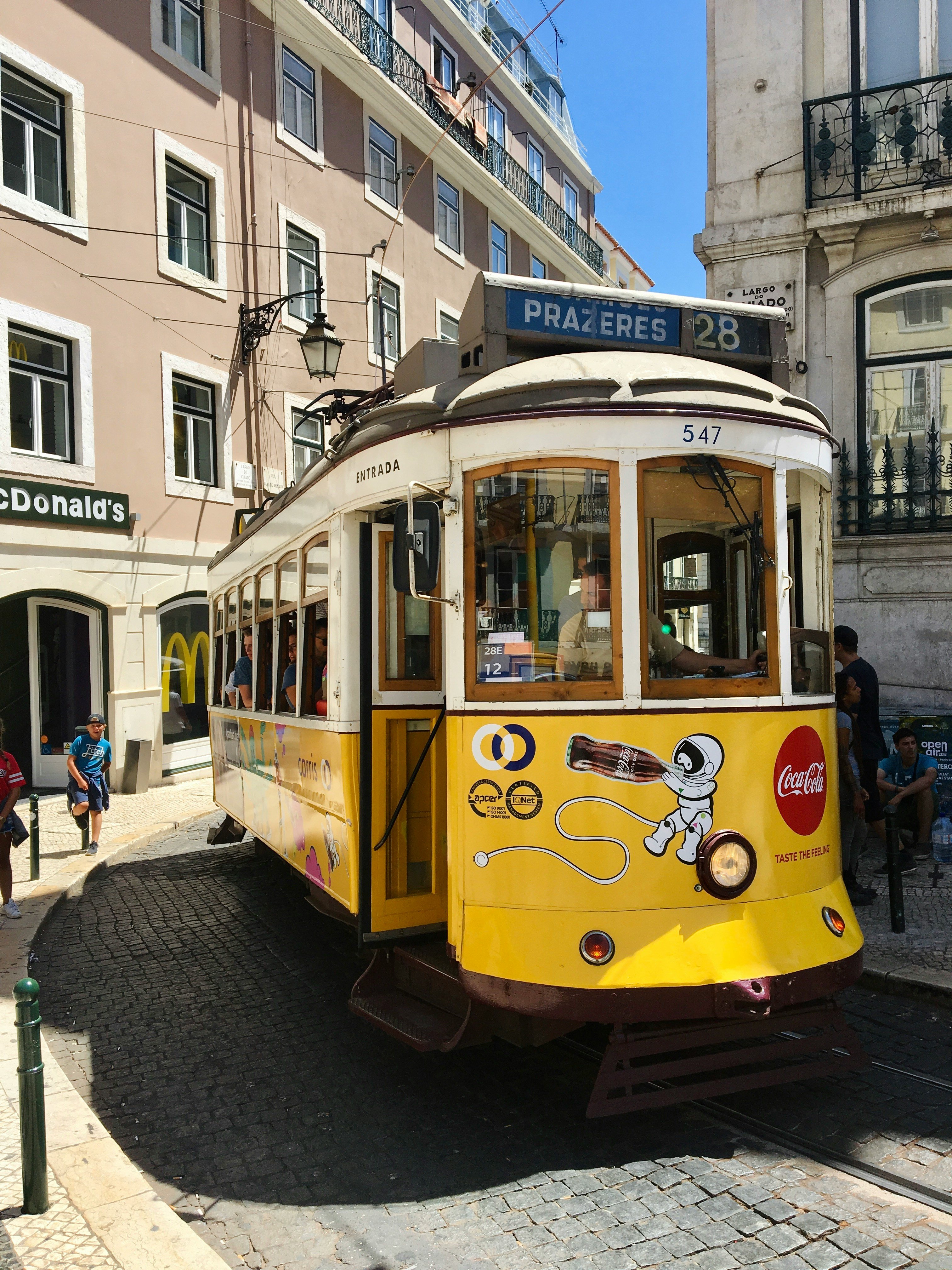 A classic yellow Tram 28 winds through the narrow, cobblestone streets of Lisbon, Portugal, passing colorful buildings and historic neighborhoods.