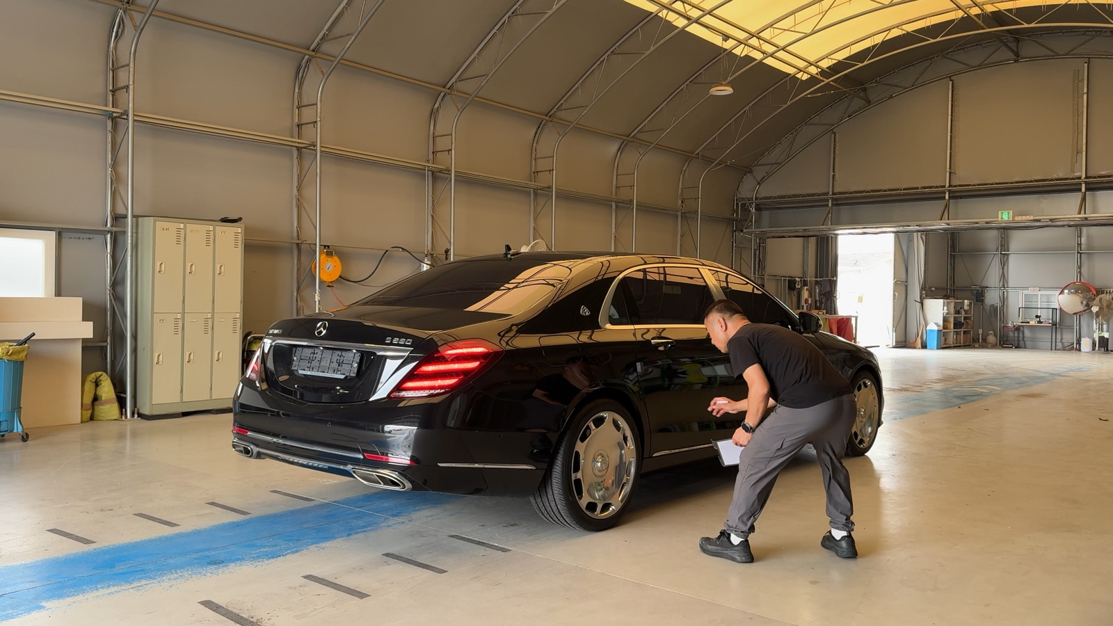 An LMN staff member with a clipboard walking around a secured black Mercedes-Maybach just after it arrived at the LMN facility, noting its condition.