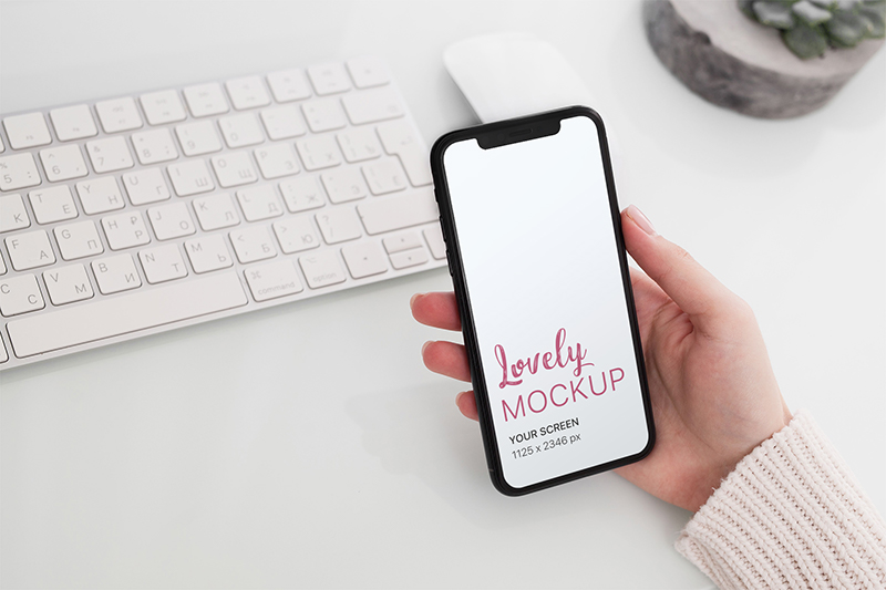 Mockup and Woman's Hand Holding an iPhone 11 over a Desk - Lovely Mockups