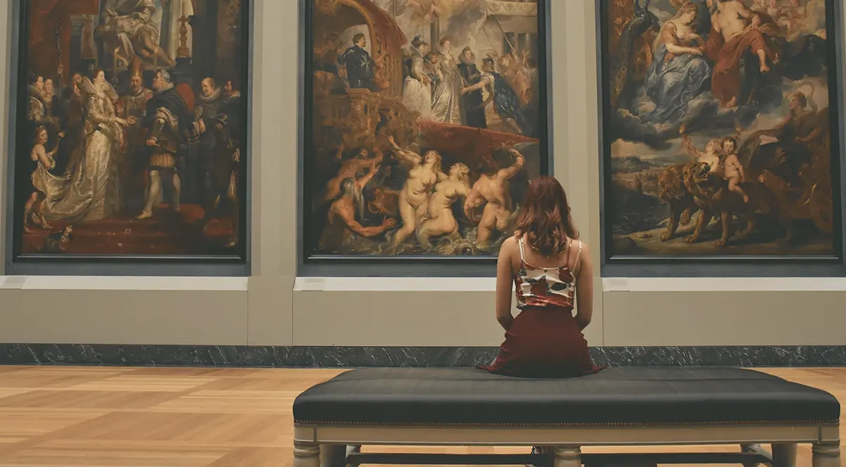 A woman enjoying a museum date while seated on a bench inside the building and gazing at three enormous paintings