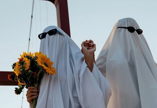 Two persons wearing a ghost costume with black sunglasses holding hands and flowers questioning why do people ghost