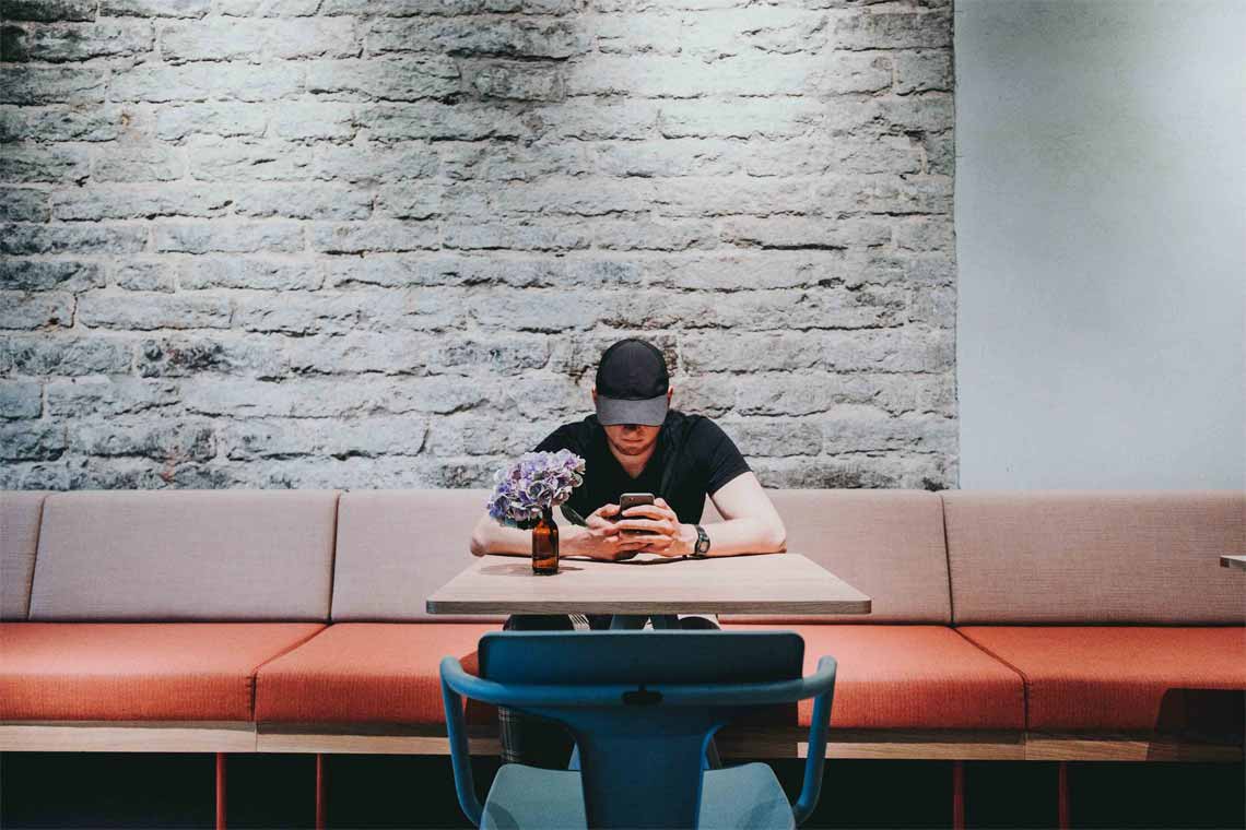 a man with cap sitting at a table while holding a cellphone