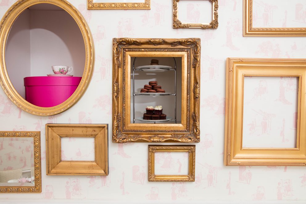 A pink wall with framed cupcakes in a gold frame.