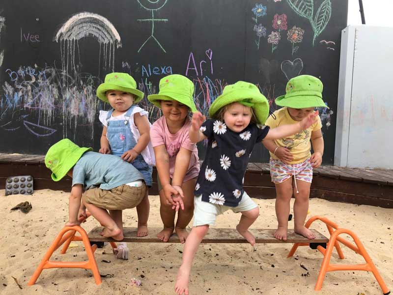 Five young children stand on a plank of wood supported by two metal horses in a sandpit. They wear green hats and summer clothes. Behind them is a black board with colourful children's drawings.