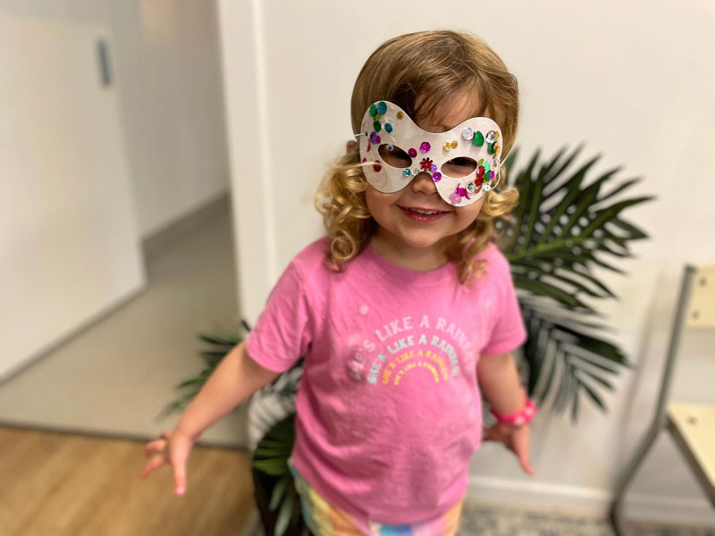 Child in a pink show that reads "Life is like a rainbow" wears a mask as she role plays.