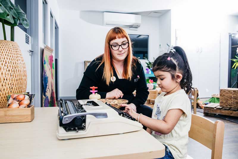 Educator observes child using an old typewriter for play-based learning while caring for children.