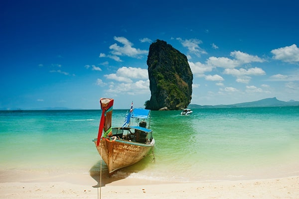 A serene beach travel package showing a traditional wooden boat resting on a calm, turquoise sea with a lush tropical island in the distance.