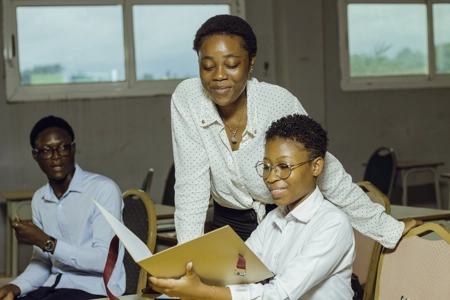Une étudiante souriante avec des lunette est assise montrant quelque chose à sa camarade en classe