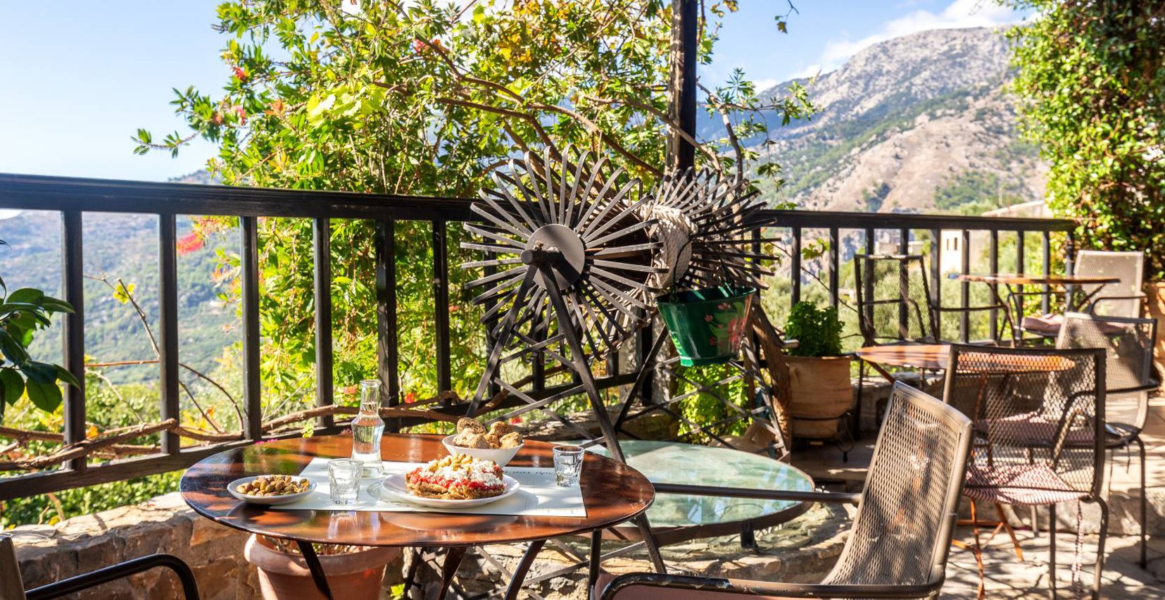 The outdoors restaurant of Mala Villa with the tables with delicious food like cretan ntakos, olives, Cretan raki, bread, the old renovated well in the side of the area, and the view of the beautiful mountains