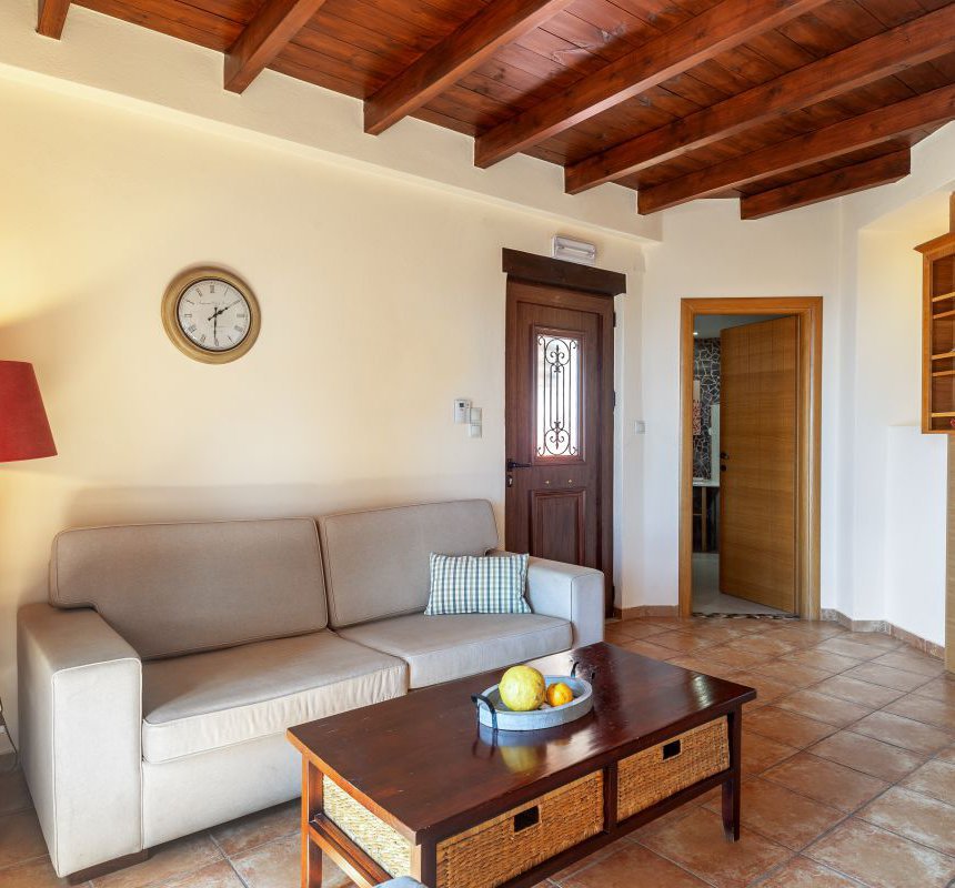 The interior of the maisonette with the modern lamp, the sofa with the wall clock above it, the coffee table with fruits on top of it, the shelves on the other side, and the wooden ceiling on top