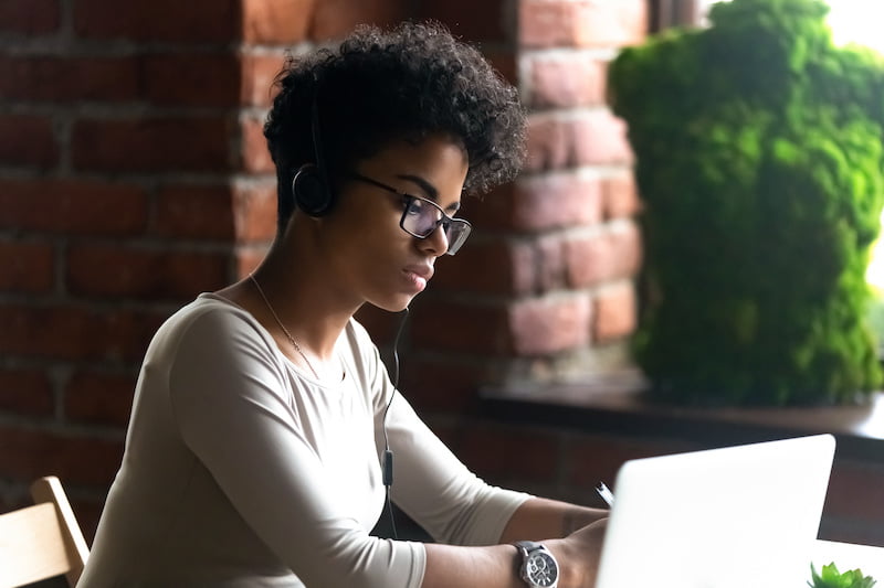 Woman wearing glasses and headphones working on laptop at brick desk