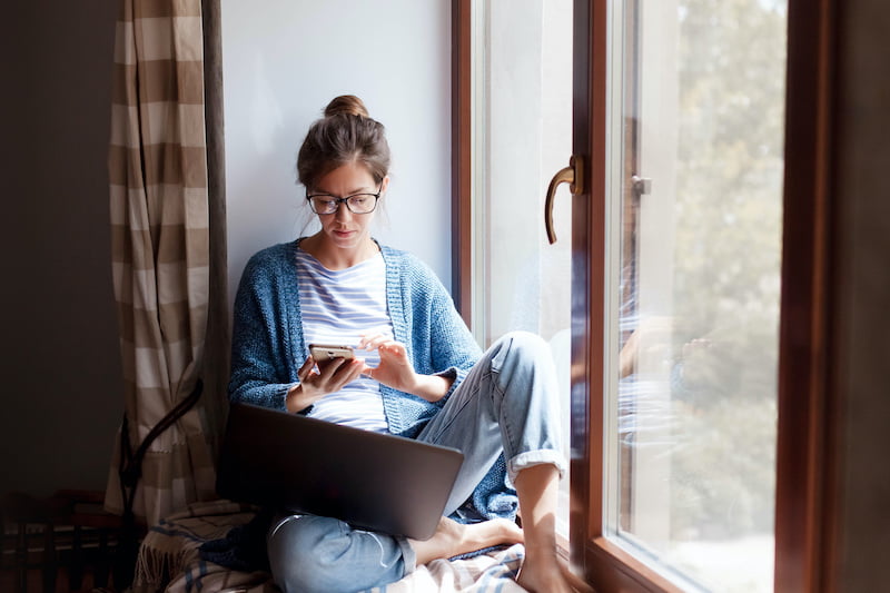 Woman with glasses working on laptop by window, holding smartphone