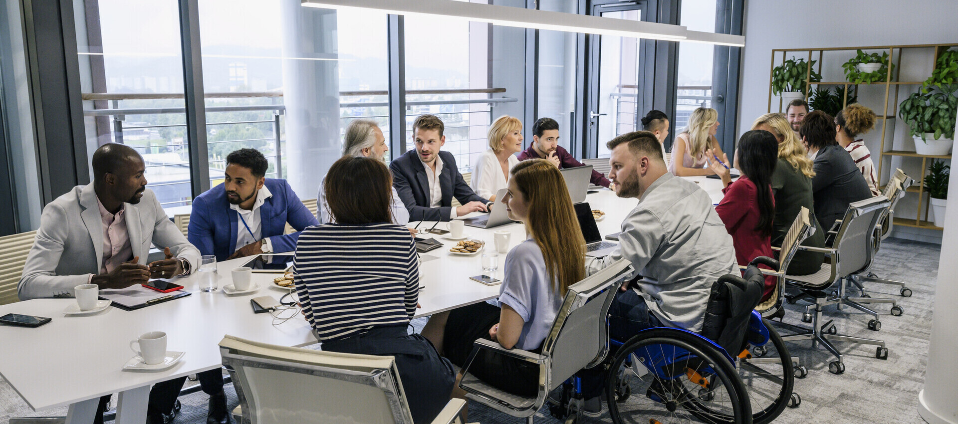 A large meeting room hosting about 15 people of diverse genders and cultures around a table. A person near the front is in a wheelchair.