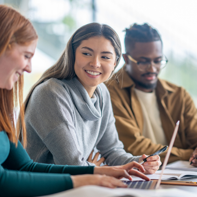 A diverse group of three young professionals collaborating in a bright, modern office or classroom setting. A woman in a gray cowl-neck sweater sits between a woman with long blonde hair in a teal top and a man wearing a tan jacket and glasses. They are leaning over a laptop and papers, appearing engaged and positive, with soft natural lighting coming through windows in the background. Their body language suggests an attentive, cooperative work environment with a sense of teamwork and shared focus.