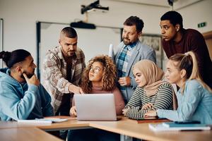 Diverse group of six young professionals gathered around a laptop in a bright, modern classroom or office space. A person in a hijab and a person in a striped sweater sit at the table while others stand, leaning in collaboratively. Warm wood tones of the desk, neutral wall colors, and natural lighting create an inviting atmosphere. Participants appear engaged, with attentive body language and a sense of teamwork. Clothing styles range from casual denim jackets to professional button-ups, suggesting an inclusive, contemporary workplace or educational setting focused on collaborative learning and interaction.