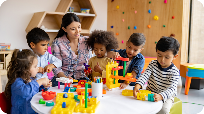 A smiling female teacher in a plaid shirt supervises five diverse young children playing with colorful building blocks at a white table.