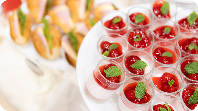 Elegant catering display with rows of strawberry panna cotta desserts garnished with mint, and blurred sandwiches in the background.