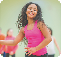 Young woman with long, curly dark hair smiling broadly while dancing or exercising, wearing a bright pink tank top. She appears energetic and joyful, moving with enthusiasm in what seems to be a fitness or dance class setting. Blurred background suggests other participants, with a soft green-tinted wall visible. Her body language conveys confidence and enjoyment of the physical activity, with an outstretched arm and dynamic posture capturing a moment of movement and expression.