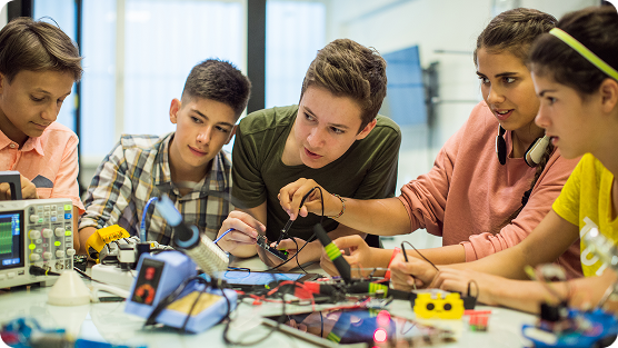 A group of young students collaborate intently in a bright, modern technology lab. Five diverse teenagers huddle around a workbench filled with electronic components, colorful wires, circuit boards, and technical measurement equipment. A blue oscilloscope and other technical devices are visible. Students wear casual clothing in shades of peach, green, yellow, and plaid, leaning closely together and focusing on hands-on technical work. The scene conveys teamwork, concentration, and hands-on learning in a STEM educational environment, with students actively engaged in exploring robotics or electronics project.