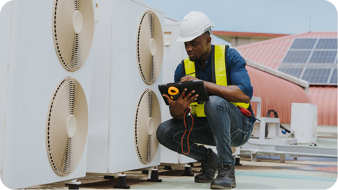 An HVAC technician wearing a hard hat and safety vest uses a digital tablet while servicing outdoor air conditioning units on a rooftop