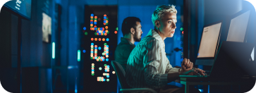 Two focused IT professionals, a woman and a man, work on laptops in a dark server room, representing the hands-on skills gained through IBM.