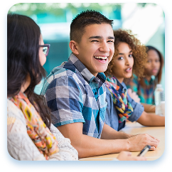 A diverse group of young students sit together at a table, engaged in conversation. A person in a blue and white plaid short-sleeve shirt smiles widely, radiating enthusiasm. Behind him, other students with varied hairstyles and clothing lean in, creating an energetic classroom or study group atmosphere. Soft teal and blurred background suggest an educational setting with natural lighting. The students appear relaxed and attentive, representing a collaborative and inclusive learning environment.
