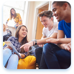 Group of diverse young people relaxing together in a casual indoor setting. A person in a blue shirt and another in a white shirt sit closely with a woman wearing a plaid shirt, all smiling and appearing engaged in friendly conversation. Another person in a yellow top is partially visible in the background. The scene looks warm and comfortable, with soft natural lighting suggesting a collaborative and social environment, like a student lounge or shared living space. Their body language and expressions convey a sense of camaraderie and positive interaction.