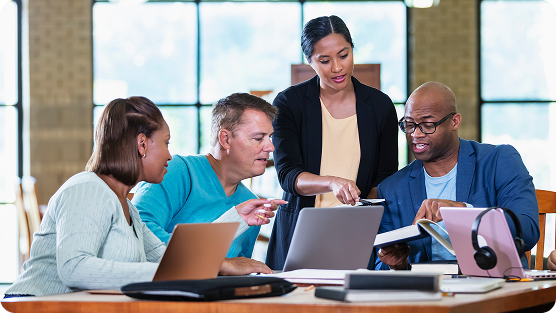 A diverse group of four professionals collaborating around a wooden table in a bright, windowed office space. A woman in a black cardigan stands, leaning over and pointing at a laptop screen while three seated colleagues - wearing teal, light blue, and navy blue tops - look on attentively. Multiple laptops, notebooks, and a pink pair of headphones are scattered across the workspace. The setting suggests a professional meeting or collaborative work session with an open, modern interior design featuring natural light and neutral tones.