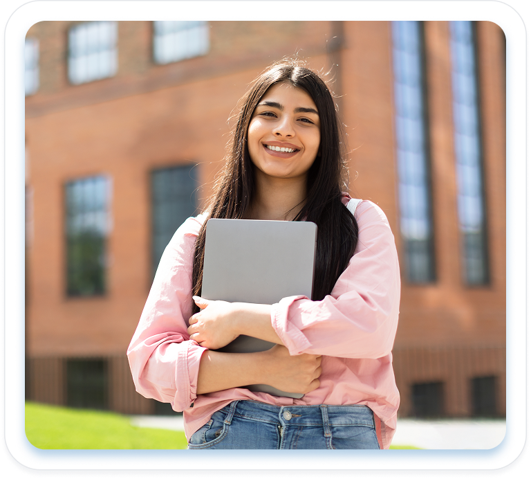 A smiling young woman with a laptop stands outside a modern university building, representing confidence and ease in the Intensive English Stream.