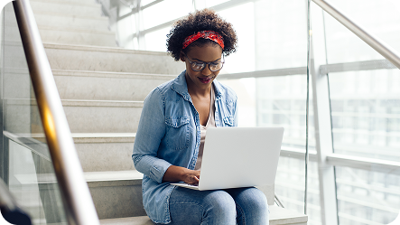A young professional sits on a white staircase with glass railings, working on a silver laptop. Wearing a denim jacket, glasses, and a red patterned headband, they have curly hair and appear focused. Soft natural light filters through nearby windows, creating a bright, modern workspace environment. The setting suggests a contemporary office or educational building with clean, minimalist architectural design.