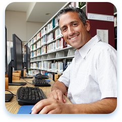 Middle-aged professional with a warm, friendly smile sits at a computer workstation in a well-lit library. He wears a crisp white short-sleeved shirt and appears comfortable and engaged. Bookshelves filled with volumes line the background, creating a scholarly environment. The workspace includes a desktop computer with a keyboard, suggesting an academic or research setting. Soft natural lighting and neutral colors contribute to a welcoming, productive atmosphere. The individual looks relaxed and confident, representing a supportive educational or professional environment.