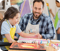 An adult and young student collaborate in a vibrant art classroom, seated at a colorful table covered with painting supplies. The student, wearing a light blue shirt, focuses intently on a creative project. The adult, dressed in a blue plaid shirt and white apron, smiles warmly while assisting the student. Painting materials, brushes, and colorful artwork are scattered across the workspace, creating an engaging and supportive learning environment with natural light and cheerful background colors.
