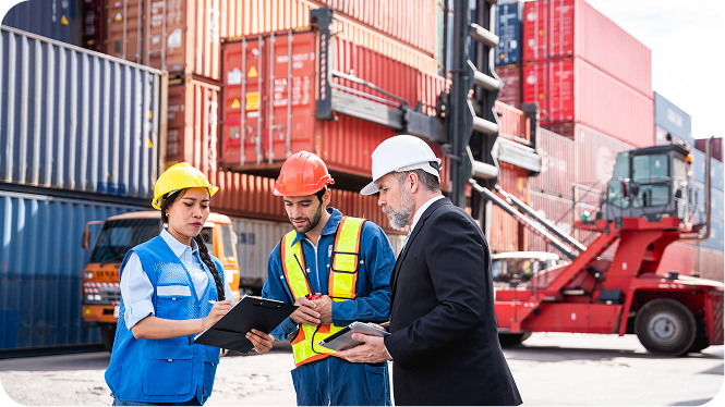 Three logistics professionals in hard hats reviewing data on a clipboard and tablet at a bustling shipping container terminal.