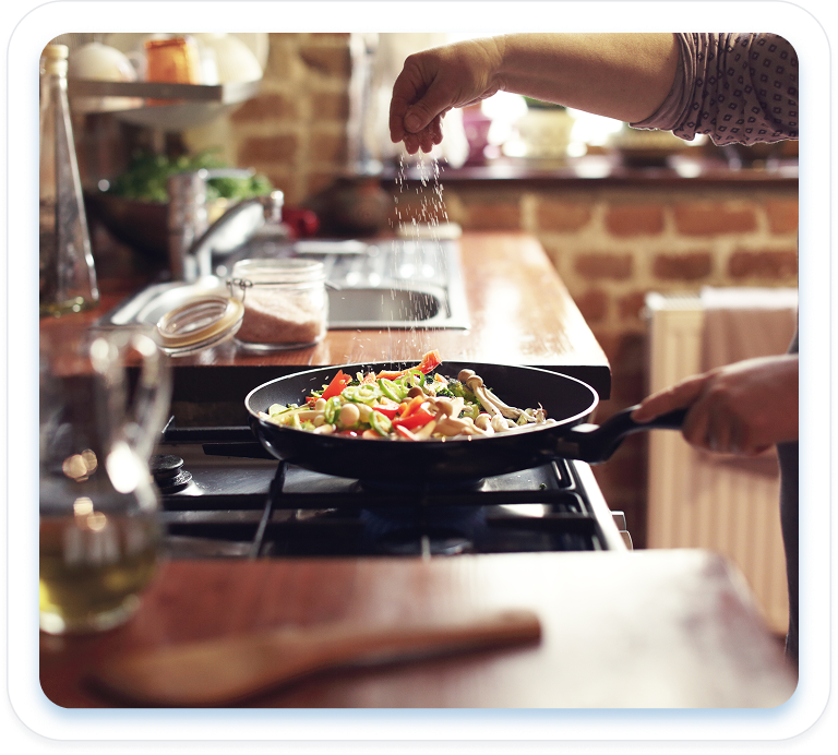 A person sprinkles seasoning into a skillet of colorful stir-fried vegetables cooking on a gas stovetop in a rustic brick kitchen.