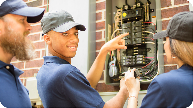 Three electrical workers in blue shirts and baseball caps inspecting an open outdoor circuit breaker panel on a brick wall. One worker points to the switches while looking toward the camera.