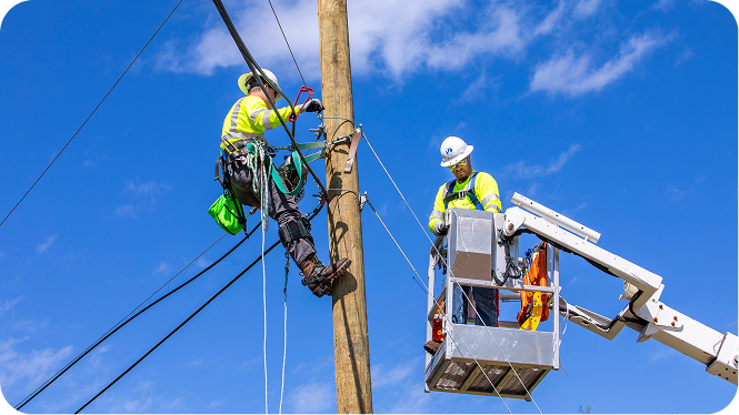 Two utility linemen wearing high-visibility gear and hard hats working on a wooden pole, one climbing with a belt harness and the other standing in a bucket lift.
