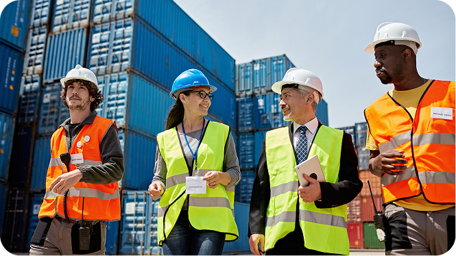 Four diverse logistics professionals wearing hard hats and high-visibility safety vests walk and talk together at a shipping port, with massive stacks of blue cargo containers in the background.