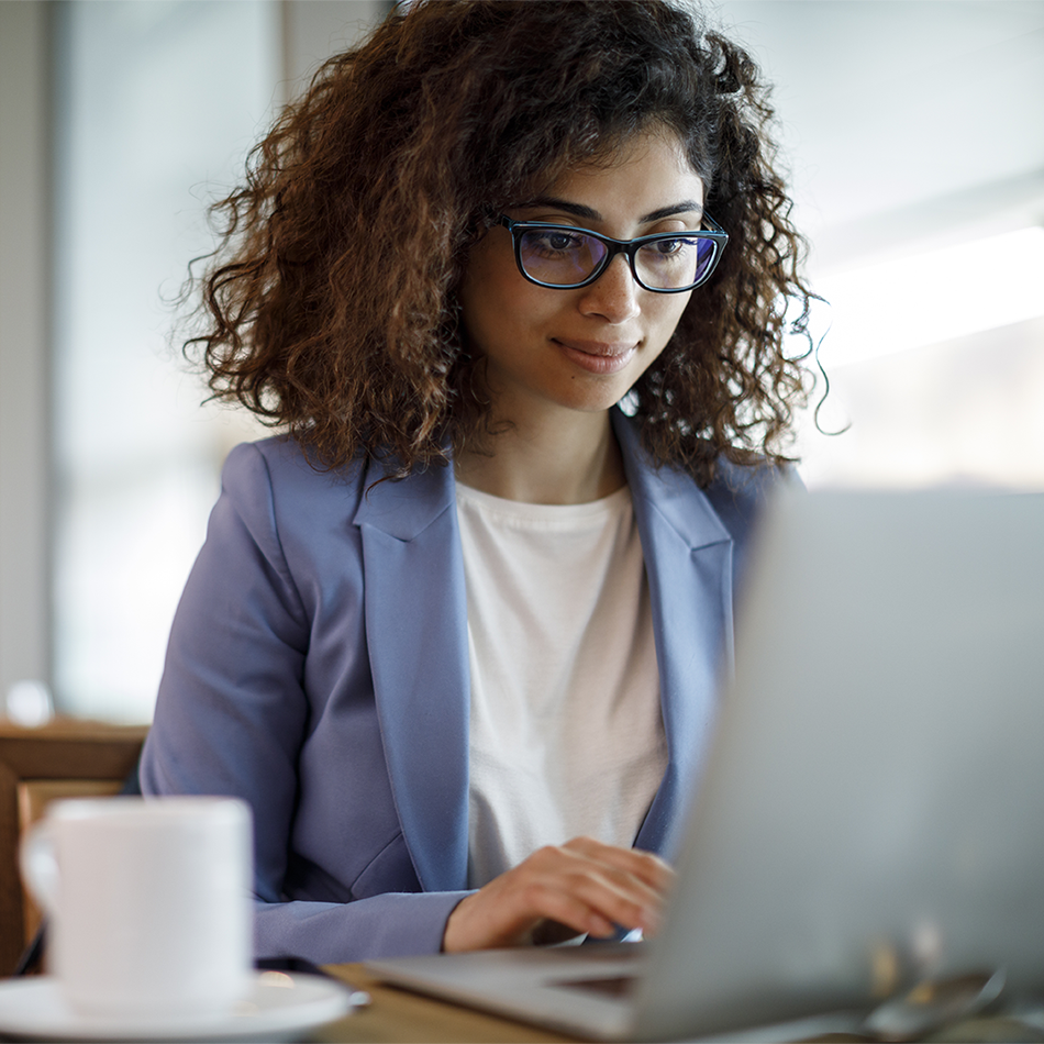 Female accounting professional with curly dark hair and blue-framed glasses wearing business blazer, focused on laptop work in modern office environment